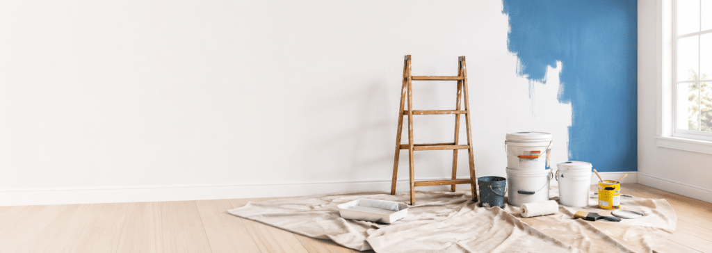 Interior wall being painted blue with ladder, paint buckets and protective floor covering
